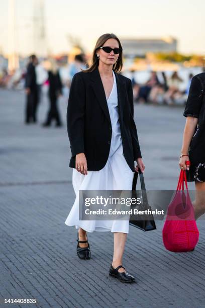 Guest wears a black sunglasses, a white V-neck / midi dress, a black blazer jacket, a black shiny leather handbag, black shiny leather slit / split...