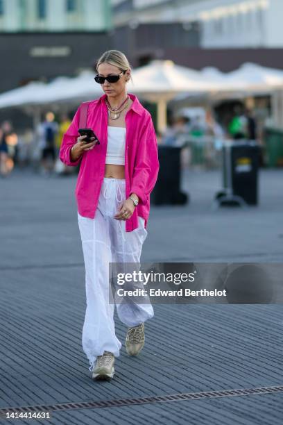 Guest wears black sunglasses from Versace, a pink with large silver pendant necklace, a gold necklace, earrings, a pink linen shirt, a white...