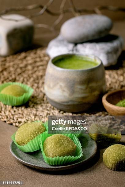 Traditional asian rice dessert sweet green matcha mochi with cup of frothed matcha tea and green matcha powder in ceramic plate on brown table....