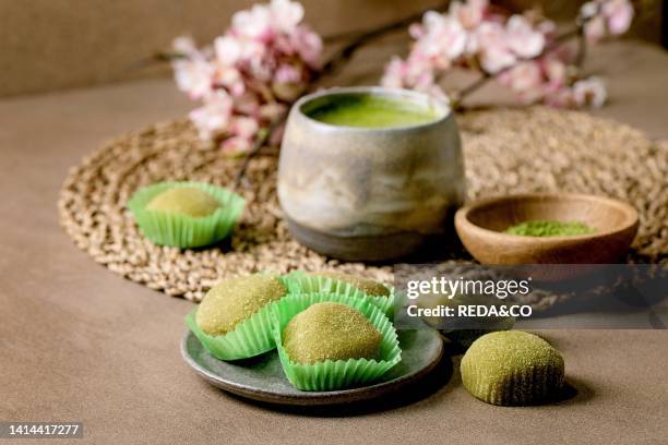 Asian rice dessert sweet green matcha mochi with cup of frothed matcha tea and green matcha powder in ceramic plate on brown table. Teapot and spring...