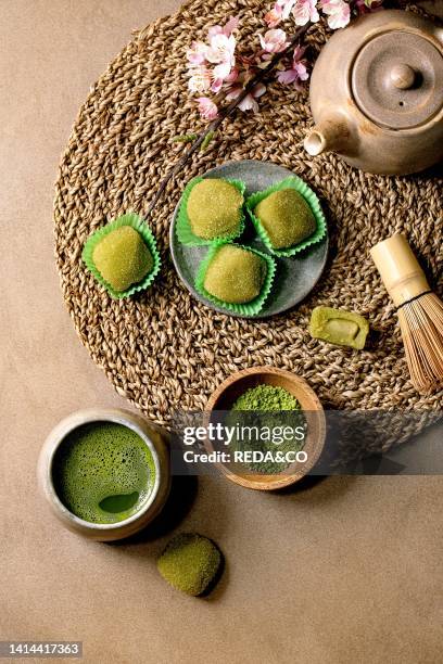 Asian rice dessert sweet green matcha mochi with cup of frothed matcha tea and green matcha powder in ceramic plate on brown table. Teapot and spring...