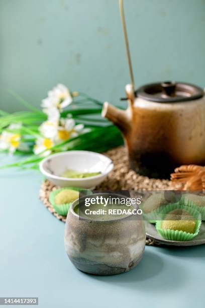 Asian traditional rice dessert sweet green matcha mochi with cup of frothed matcha tea on blue table. Old teapot and spring daffodils flowers at...