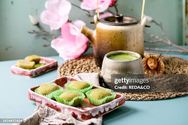 Asian traditional rice dessert sweet green matcha mochi in pink plate with cup of frothed matcha tea on blue table. Old teapot and spring pink...