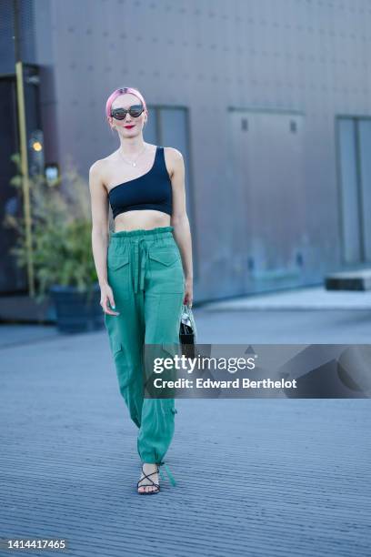 Guest wears black sunglasses, a gold chain pendant necklace, a black asymmetric shoulder strap cropped top, high waist green linen cargo pants, a...