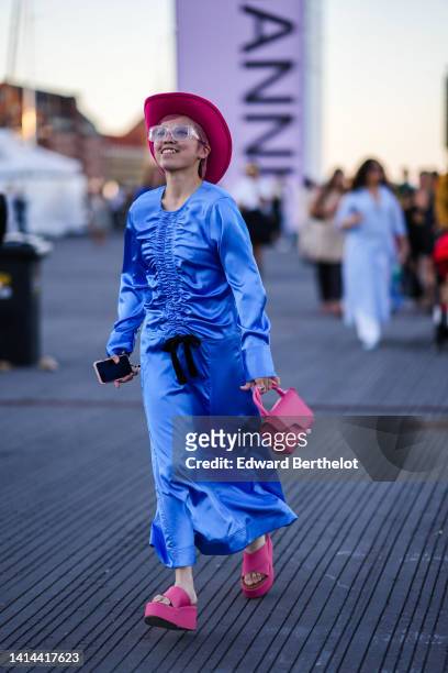 Guest wears a neon pink felt hat, glasses, a royal blue shiny satin / silk ruffled / long sleeves long dress with a black knot belt, a pink matte...
