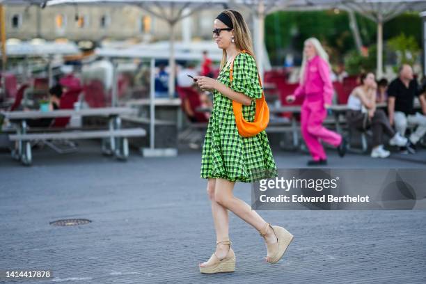 Guest wears sunglasses, a neon green and dark green checkered print pattern puffy long sleeves short dress, an orange shiny leather shoulder bag,...