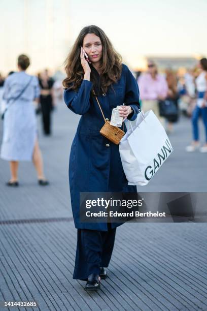 Guest wears a navy blue linen long jacket, navy blue large pants, a beige brown wicker crossbody bag, a white shopping bag, black shiny leather...