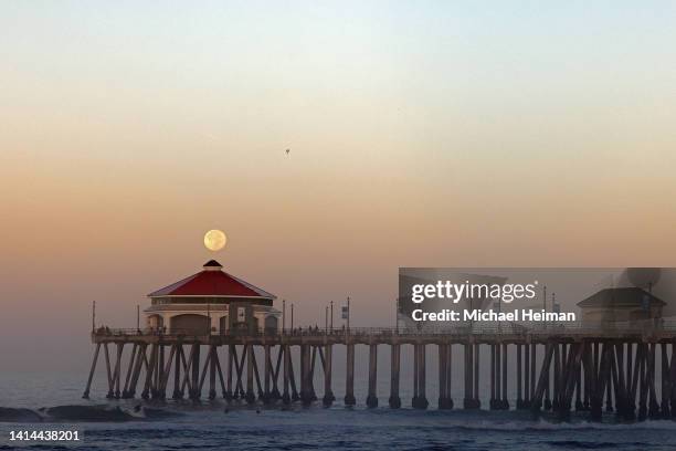 Surfer catches a wave as the Sturgeon Moon sets behind the Huntington Beach Pier over the Pacific Ocean on August 12, 2022 in Huntington Beach,...