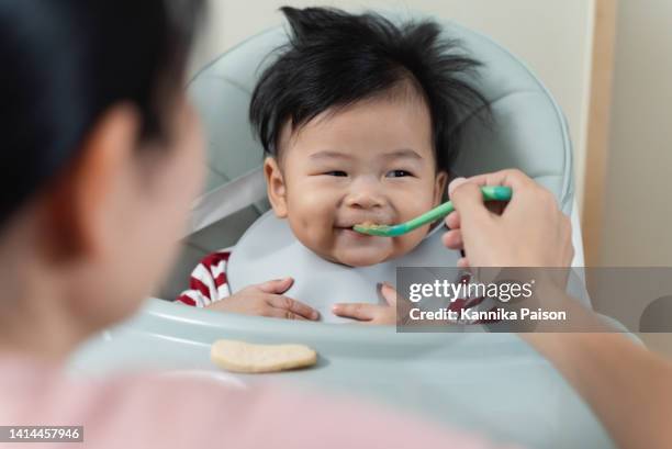 little cute asian baby boy in high chair being fed food by mom. - asian spoon feeding happy stock pictures, royalty-free photos & images