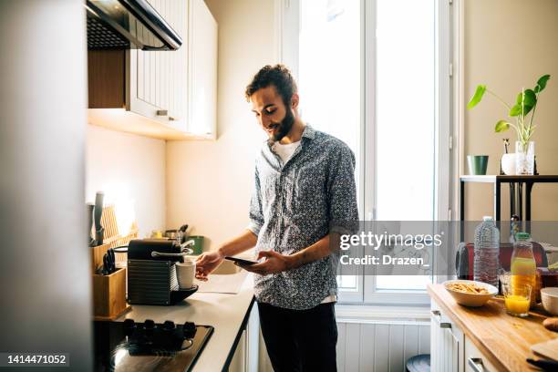 middle eastern man preparing coffee on coffee machine in modern kitchen and using smart phone - koffiemachine stockfoto's en -beelden