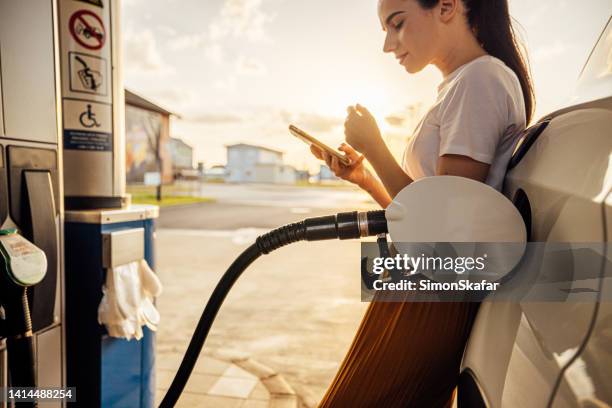 giovane donna che usa il telefono cellulare mentre fa rifornimento alla sua auto alla stazione di servizio - fare il pieno di benzina foto e immagini stock