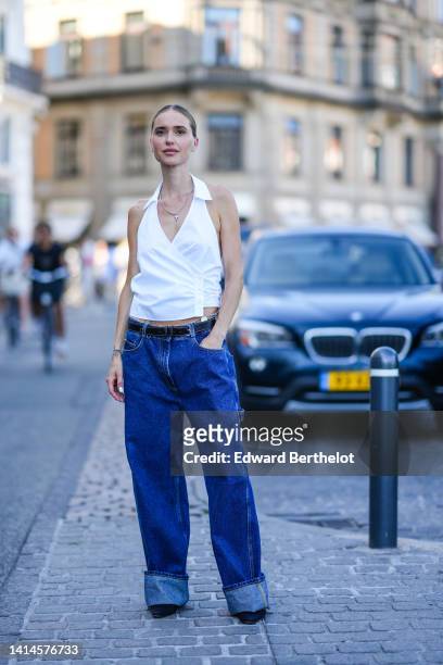 Pernille Teisbaek wears a silver chain with a cross pendant necklace, a white sleeveless / wrap gilet, a black shiny leather belt, navy blue denim /...
