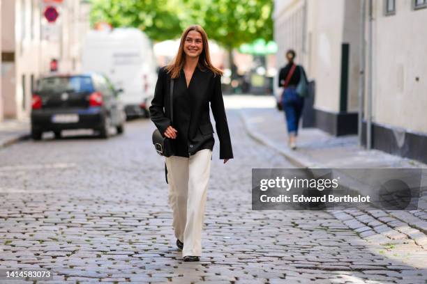 Sophia Roe wears a black blazer jacket, a black matte leather with a silver large buckle shoulder bag, white latte wide legs pants, black shiny...