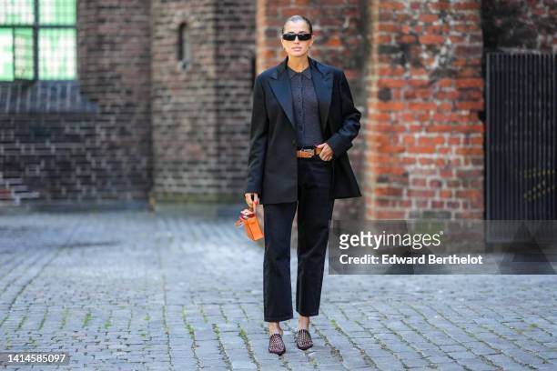 Darja Barannik wears black sunglasses, gold earrings, a dark gray wool polo shirt, a black oversized blazer jacket, an orange shiny leather Mini...