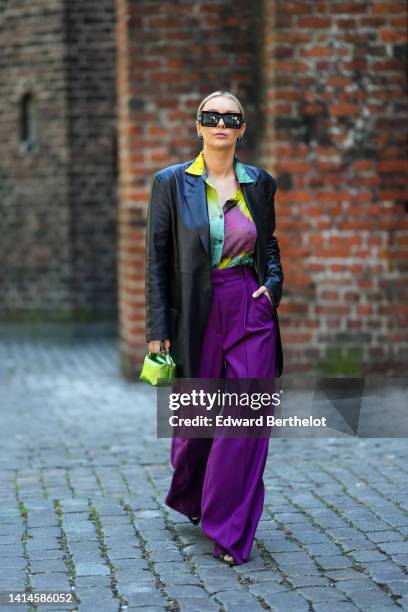 Justyna Czerniak wears black square sunglasses, a black shiny leather long blazer jacket, a purple / green / yellow tie and dye print pattern silk...