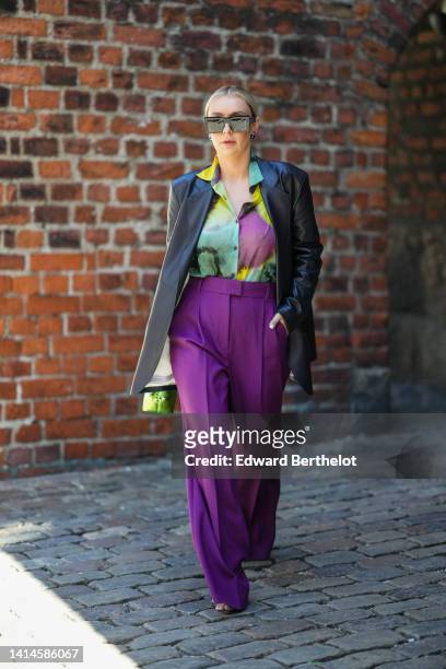 Justyna Czerniak wears black square sunglasses, a black shiny leather long blazer jacket, a purple / green / yellow tie and dye print pattern silk...
