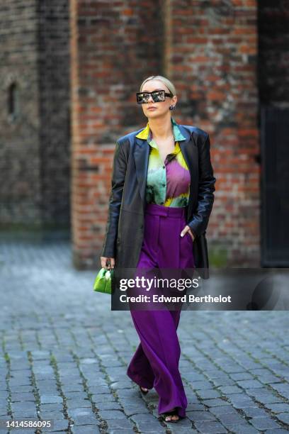 Justyna Czerniak wears black square sunglasses, a black shiny leather long blazer jacket, a purple / green / yellow tie and dye print pattern silk...