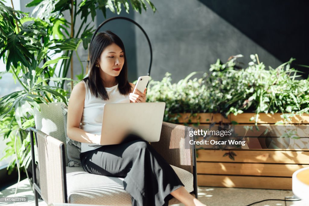 Young woman using smart phone while working on laptop in co-working space