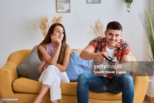 woman looking bored while her boyfriend playing video games sitting on a sofa at home. - bored girlfriend stock pictures, royalty-free photos & images