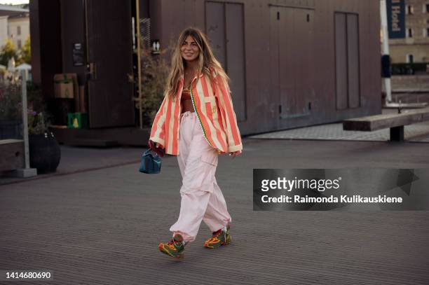 Guest wearing light pink cargo pants, orange bikini top, orange and pale yellow striped shirt, yellow, red and green sneakers posing outside Ganni...