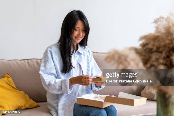 woman receiving subscription box. monthly delivery service concept. asian female looking excited at craft paper wrapped products at living room. unleashing small containers with beauty and skin products. showing positive emotion, horizontal - korean beauty products foto e immagini stock