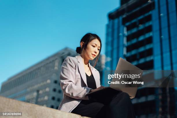 young businesswoman working on laptop, sitting against financial buildings in the city - financial occupation stock-fotos und bilder