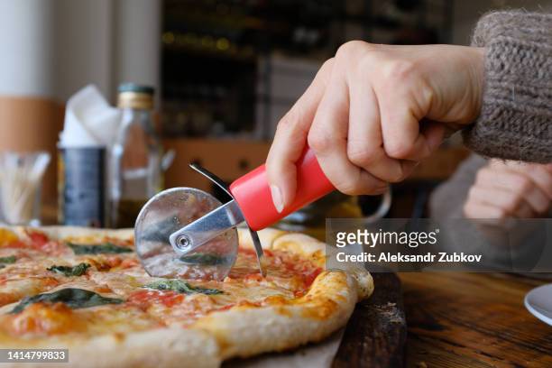 a woman cuts a piece of vegetarian pizza with mozzarella cheese, tomatoes, basil in a cafe or fast food restaurant, in a pizzeria. delicious italian cuisine. sliced pizza margarita on a wooden board or stand for serving dishes. unhealthy lunch, snack. - ピザカッター ストックフォトと画像
