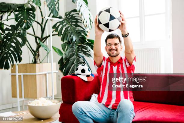 young man cheering while watching tv at home - red sports jersey stock pictures, royalty-free photos & images