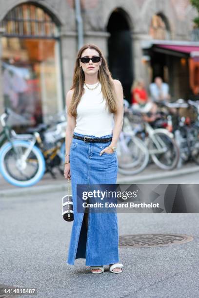 Annabel Rosendahl wears black sunglasses, earrings, a gold large chain necklace, a white ribbed halter-neck tank-top, a black braided shiny leather...