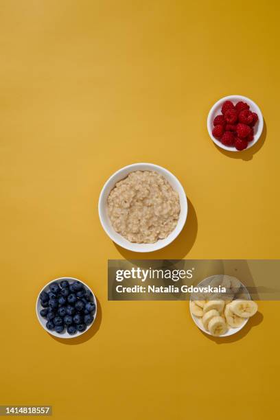 oatmeal with blueberry, raspberry and banana on yellow background. morning breakfast served in white bowl. flatlay group of objects. sweet vegan porridge recipe. showing cooking process. vertical, above, copy space - haferflocken stock-fotos und bilder