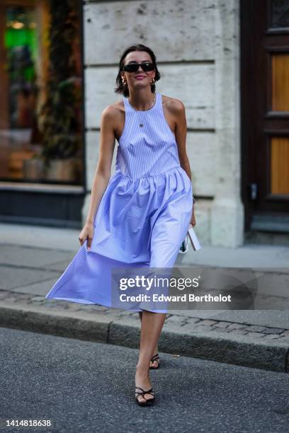 Guest wears black sunglasses, gold earrings, a gold chain pendant necklace, a baby blue and purple striped print pattern sleeveless / ruffled midi...