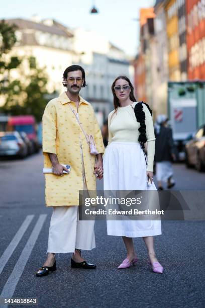 Guest wears sunglasses, silver earrings, a white t-shirt, a pale yellow with embroidered flower pattern long coat, white matte ripped wide legs...