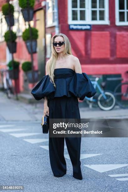 Guest wears black sunglasses, a black shoulder-off / oversized puffy short sleeves / jumpsuit, a black shiny leather handbag, pumps heels shoes ,...