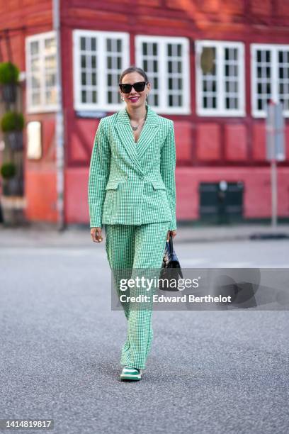 Guest wears black sunglasses, silver earrings, a silver chain pendant necklace, a white and green checkered print pattern blazer jacket, matching...