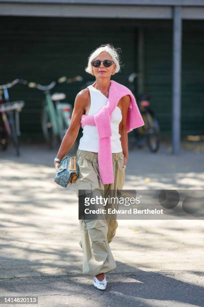 Grece Ganhem wears black sunglasses, a white ribbed tank-top, a pale pink wool knot shoulder pullover, a silver watch from Rolex, a blue denim...