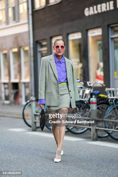Guest wears white and red sunglasses, silver and rhinestones pendant earrings, a silver chain necklace, a neon purple shirt, a beige and brown small...
