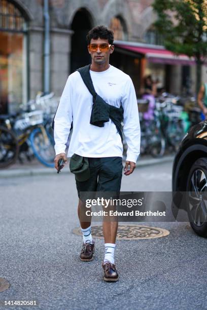 Guest wears orange sunglasses, a black knot sweater, a white long sleeves t-shirt, black shorts, a dark green cap, white socks, dark brown shiny...