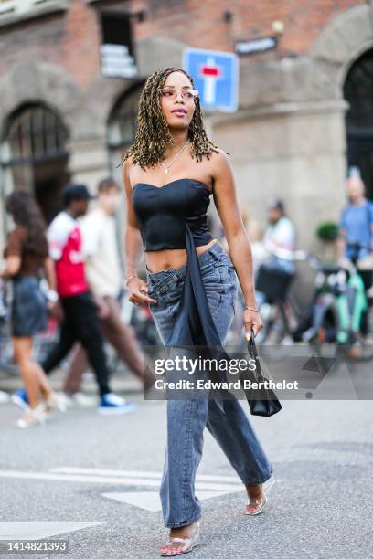 Ellie Delphine wears glasses, a gold chain pendant necklace, a white pearls necklace, a black shoulder-off / cropped top with an embroidered long...