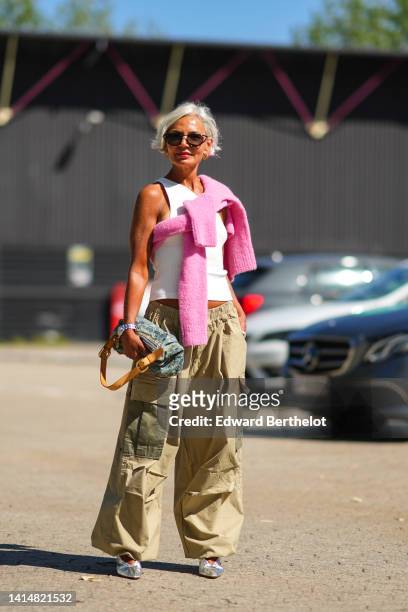 Grece Ganhem wears black sunglasses, a white ribbed tank-top, a pale pink wool knot shoulder pullover, a silver watch from Rolex, a blue denim...
