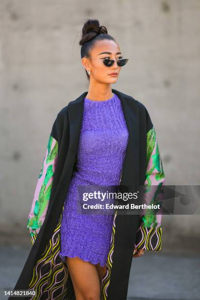 Nephtys Laurent wears black cat eyes sunglasses, silver earrings, a neon purple ruffled short dress, a black long felt with pink and green print...