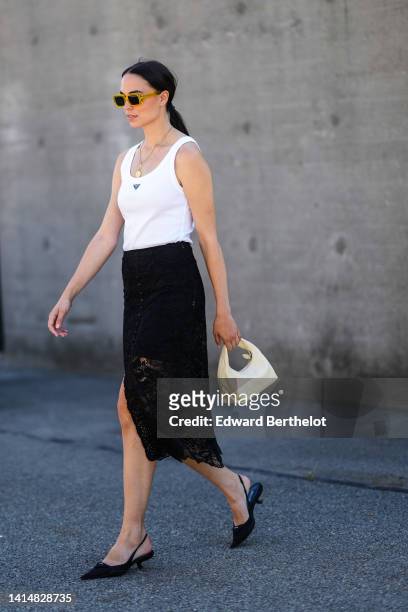 Guest wears yellow sunglasses, a silver long chain pendant necklace, a white tank-top from Prada, a black lace flower print pattern midi skirt, a...