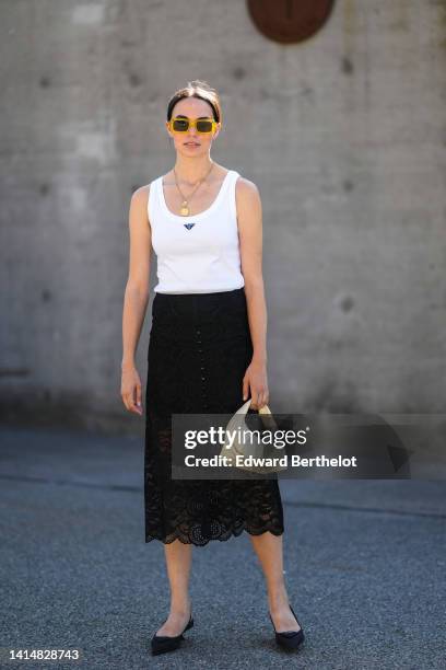 Guest wears yellow sunglasses, a silver long chain pendant necklace, a white tank-top from Prada, a black lace flower print pattern midi skirt, a...