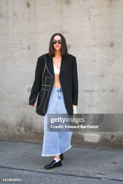 Guest wears black sunglasses, a white V-neck cropped tank-top, a black oversized blazer jacket, blue faded denim ripped long skirt, a beige matte...