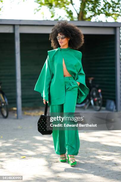 Guest wears futurist sunglasses, a green high neck / long sleeves / open blouse matching green suit pants, a silver pearls bra underwear, a black...