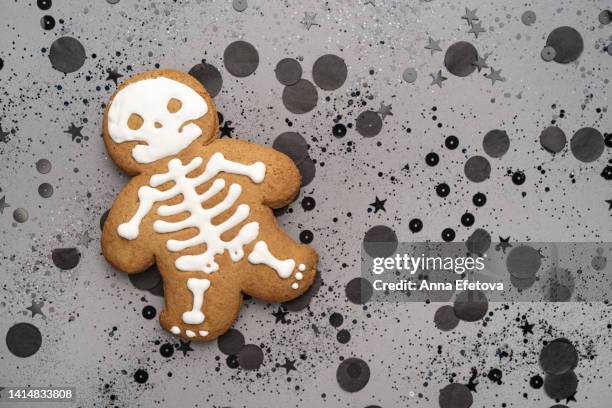 skeleton patterned gingerbread cookie placed on many small glittering round and star shaped confetti on gray background. festive halloween backdrop. flat lay style - süßgebäck teilchen stock-fotos und bilder