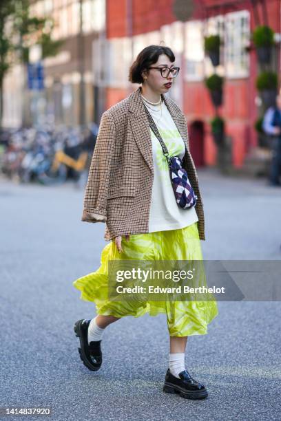 Guest wears black glasses, a white with green inscriptions print pattern oversized t-shirt, a brown checkered print pattern blazer jacket, a yellow...