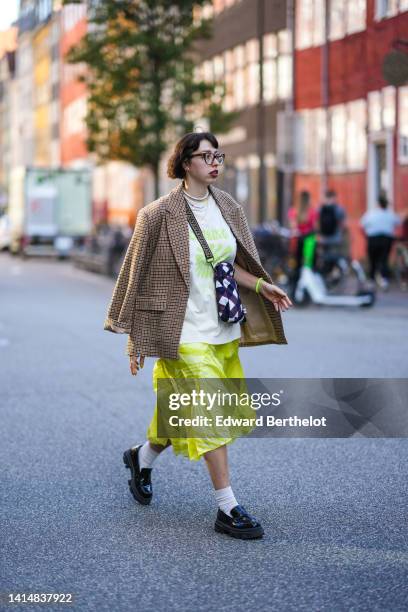 Guest wears black glasses, a white with green inscriptions print pattern oversized t-shirt, a brown checkered print pattern blazer jacket, a yellow...