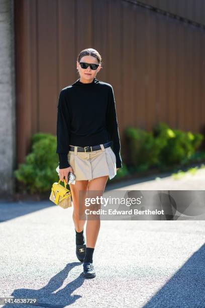 Guest wears black sunglasses, a black high neck pullover, a black shiny leather belt, a beige pleated / ripped short skirt from Miu Miu, a beige...