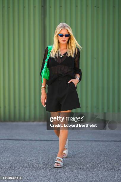 Guest wears black sunglasses, a black bra underwear, a black transparent / pleated long sleeves blouse, black shorts, a neon green shiny leather...