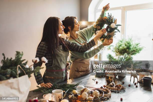 two women making christmas wreath using fresh pine branches and festive decorations. - blumenschmuck stock-fotos und bilder